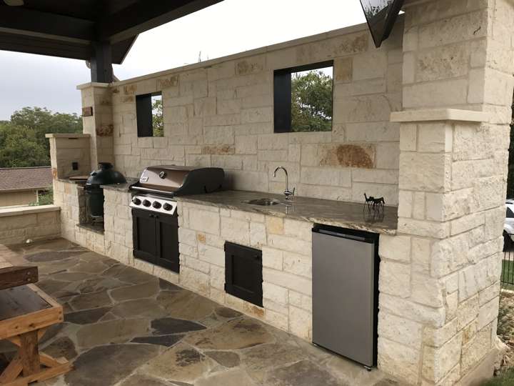 Outdoor kitchen in chopped limestone with grill, sink and refrigerator under a covered patio
