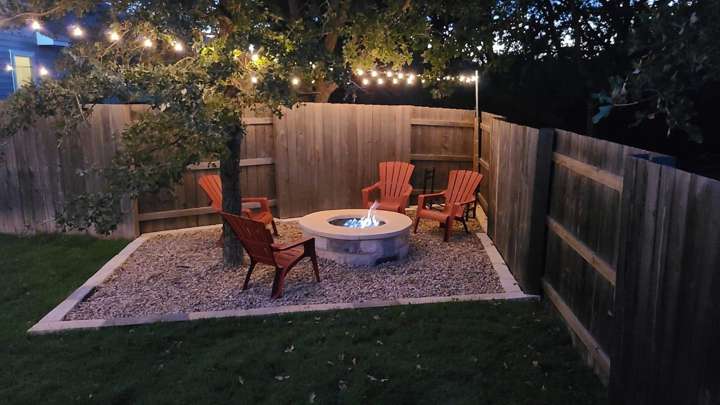 Round stone gas fire pit on a gravel pad with four chairs under string lights