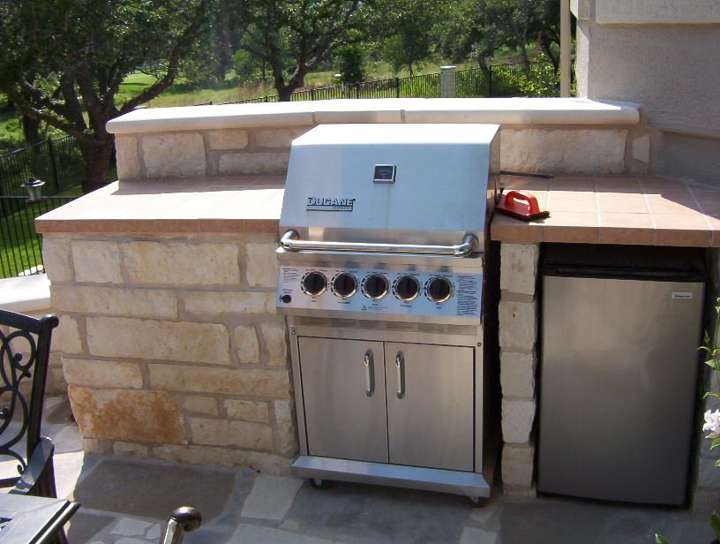 Limestone outdoor kitchen with grill and mini-fridge beside a covered porch