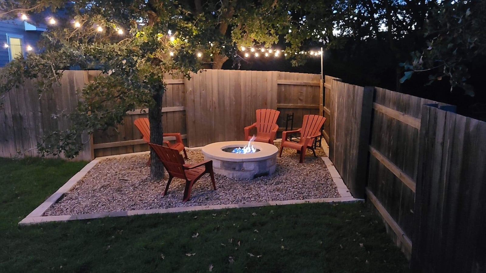 Round stone gas fire pit on a gravel pad with four chairs under string lights against a wood fence, a B&B project in Williamson County, Texas